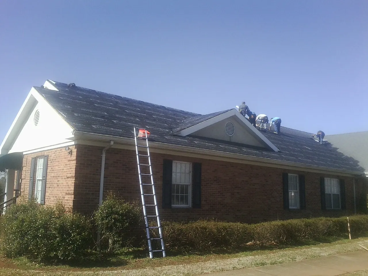 Crew tearing shingles off a brick ranch roof with a ladder set — Metal Roof Repair work by our crew in Knierim