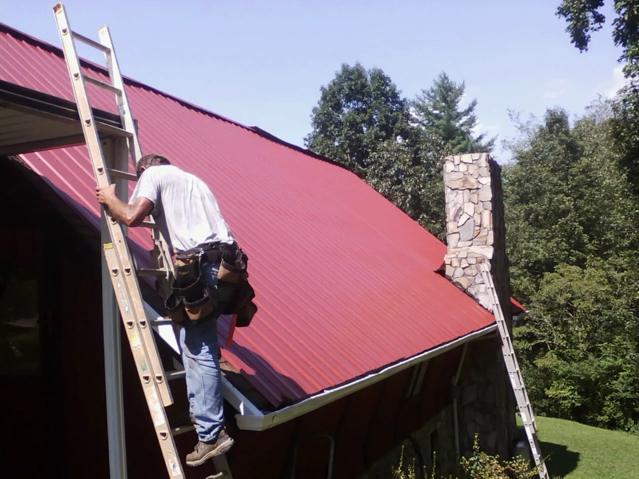 Roofer on a ladder beside a red metal roof and stone chimney — Soffit & Fascia Repair by Roofing Brothers in Knierim